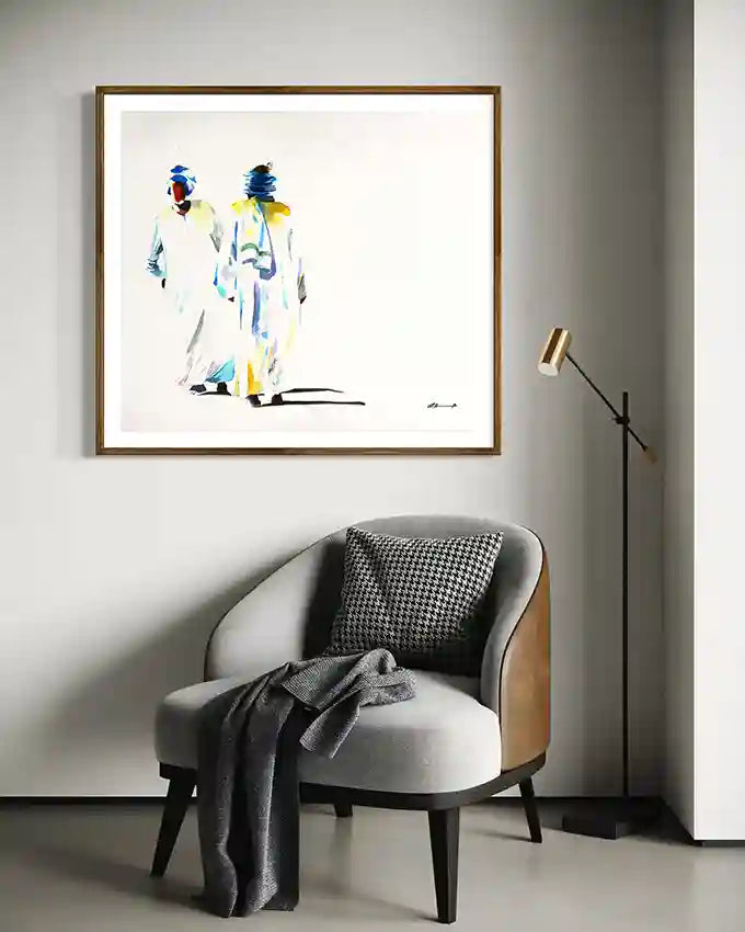 Framed art print by Sudanese Visual artist Galal Yousif on a wall above a chair with a gray blanket and patterned pillow.