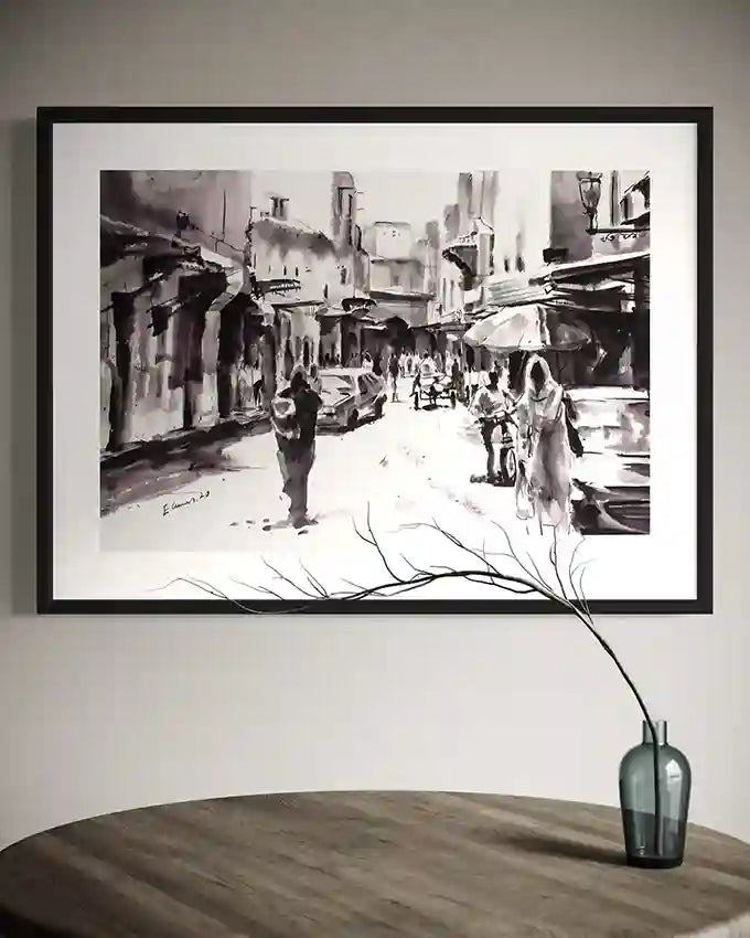 Framed black and white street scene artwork by Elamin Awad hanging above wooden table with glass vase and branch