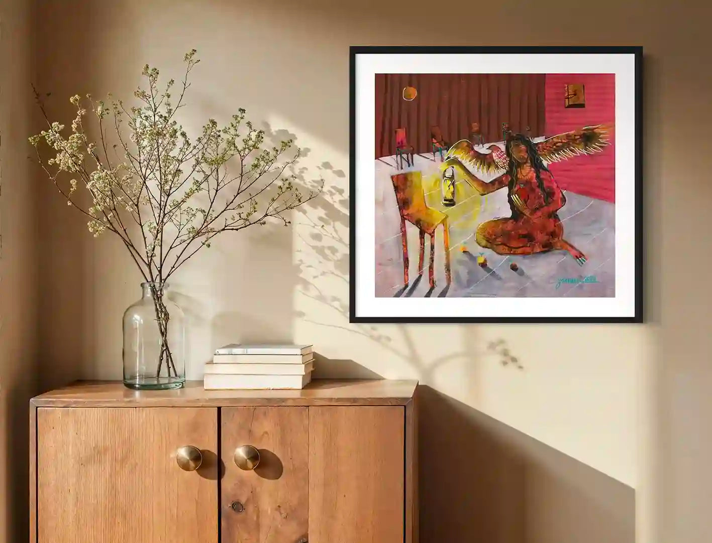 A Free Thought art print by Yasmeen Abdullah displayed in black frame above wooden cabinet with glass vase and books