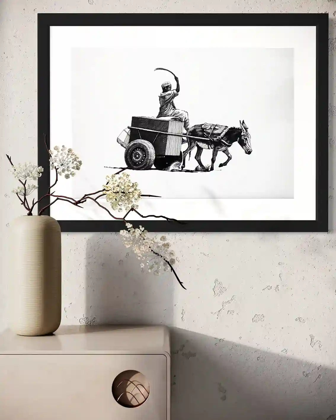 Echoes of the Road framed ink painting by Obada Gabir displayed on wall above white console with ceramic vase and flowers